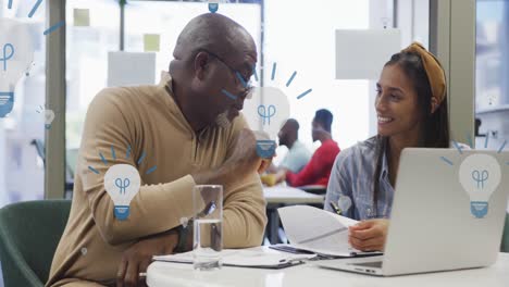 Animation-of-multiple-bulb-icons-floating-against-diverse-man-and-woman-discussing-at-office