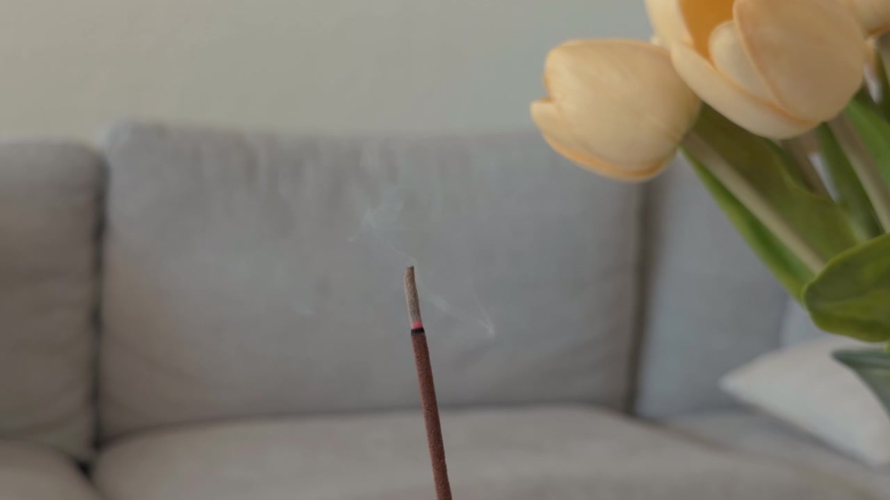 Gros plan fixe de fumée d'encens s'écoulant près de fleurs et d'un canapé à l'intérieur.