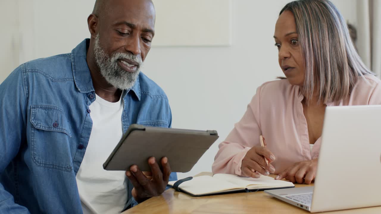 Senior couple reviewing tablet prompt, man holding tablet pointing, woman taking notes at table