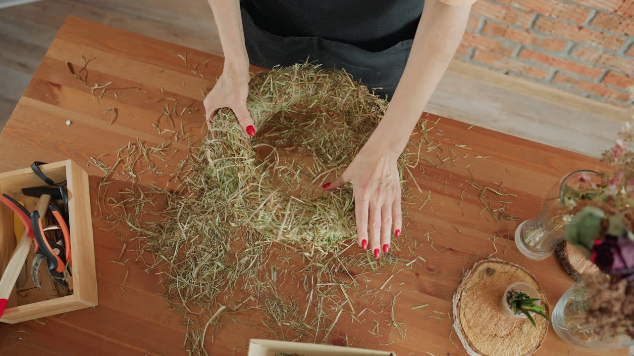 Woman holding handmade straw wreath on wooden table with scattered hay, craft tools, and natural decor elements, showcasing rustic creative decoration process atmosphere in indoor workshop