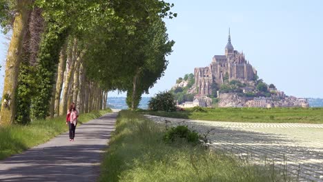 a woman walks on a pretty road through the countryside with mont saint michel monastery island in distance normandy france