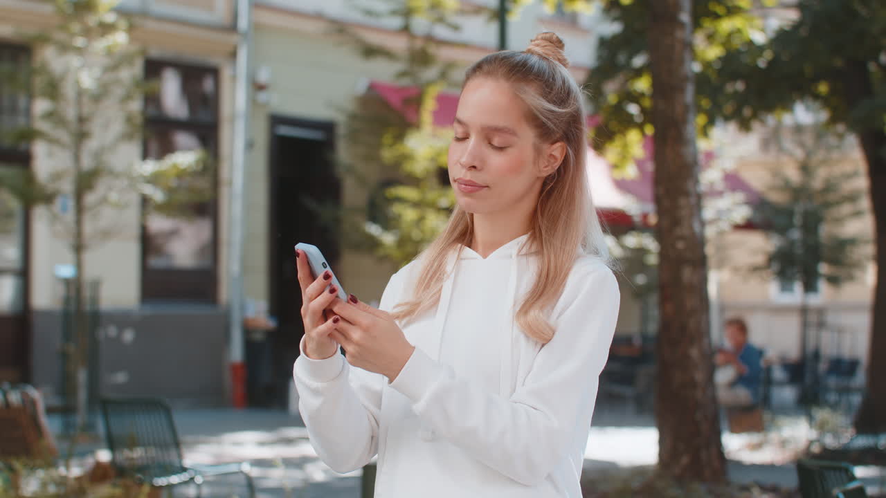 Smiling caucasian young woman using smartphone typing text messages and looking at camera in city