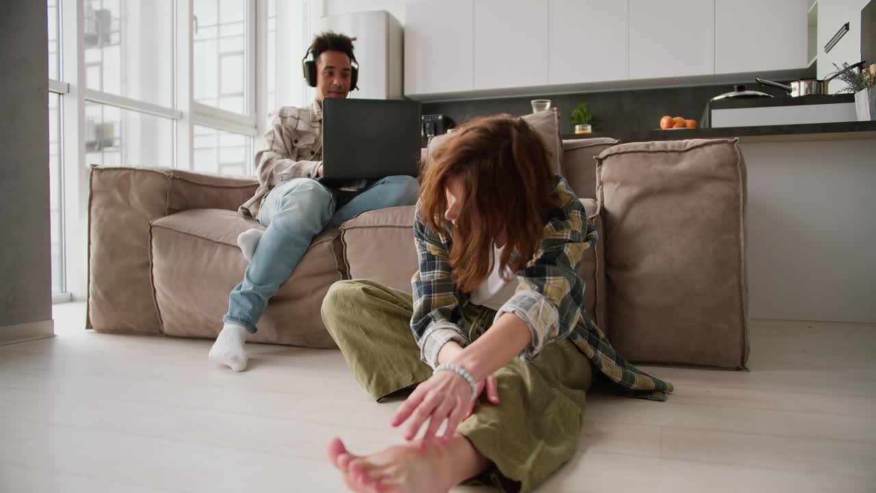 A young adult girl with brown hair in a green plaid shirt sits on the floor near the sofa and reaches for her bare feet during a yoga class while her young Black brunette boyfriend works on a gray laptop in a modern apartment