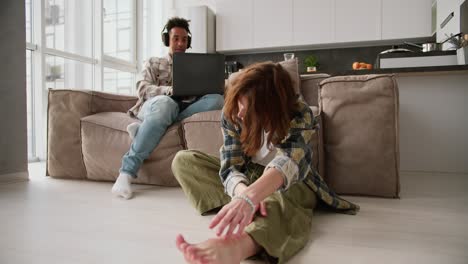 A-young-adult-girl-with-brown-hair-in-a-green-plaid-shirt-sits-on-the-floor-near-the-sofa-and-reaches-for-her-bare-feet-during-a-yoga-class-while-her-young-Black-brunette-boyfriend-works-on-a-gray-laptop-in-a-modern-apartment