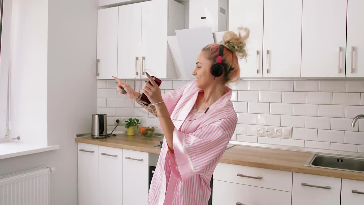 Giovane donna felice con i capelli rosa che balla in cucina, indossando vestiti rosa e ascoltando musica con le cuffie