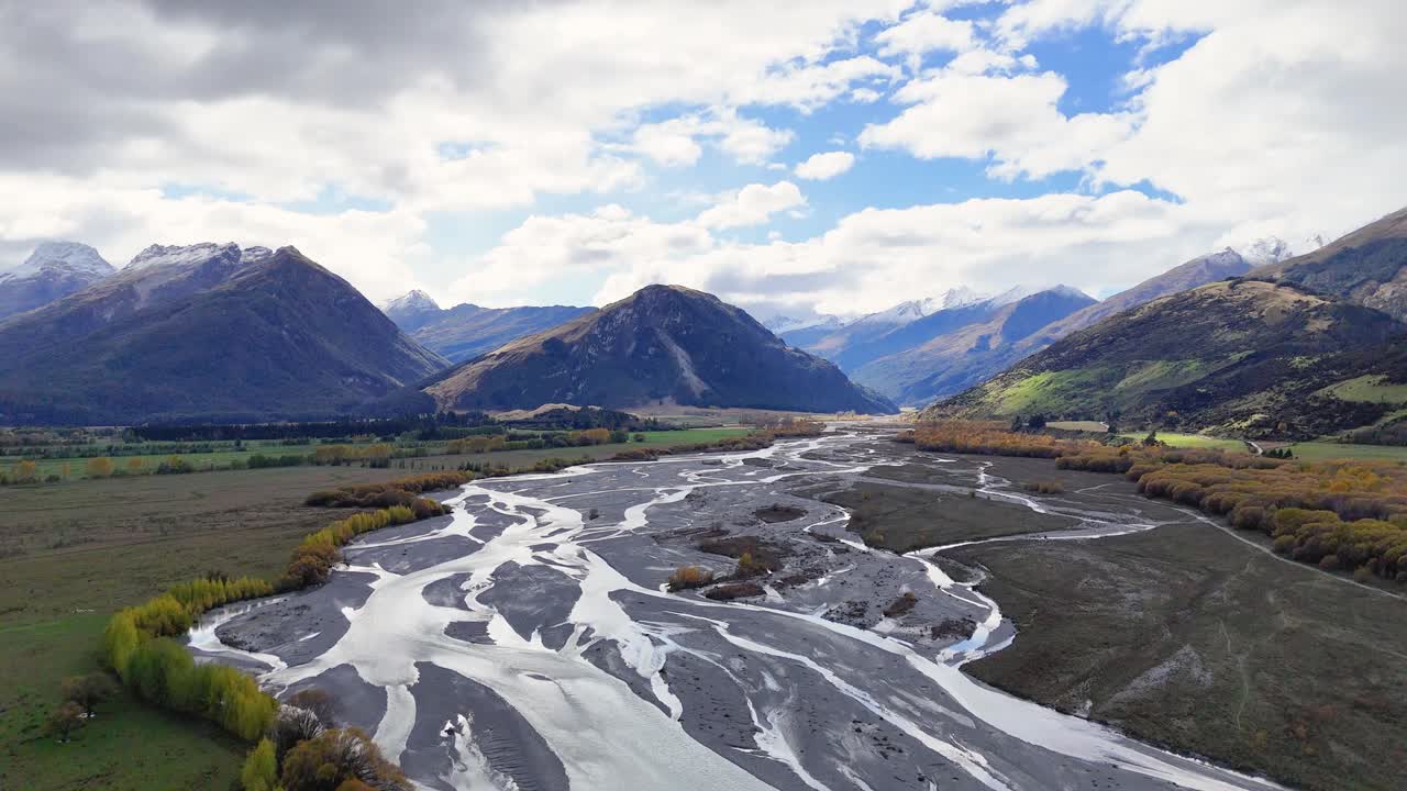 Aerial footage captures Glenorchy's braided river, surrounded by mountains under dynamic cloud cover and sunlight, creating a serene natural scene