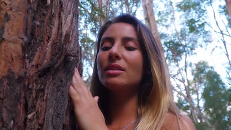 young woman touches tree bark in the forest - girl looking up while touching the rough bark of tree