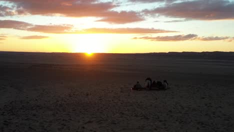 Toma-Aérea-Del-Atardecer-Del-Desierto-Sobre-Un-Grupo-De-Personas-En-Las-Dunas-De-Arena-Ondulada-En-Silueta-Seguida-Por-La-Luz-Del-Sol-En-La-Hora-Dorada