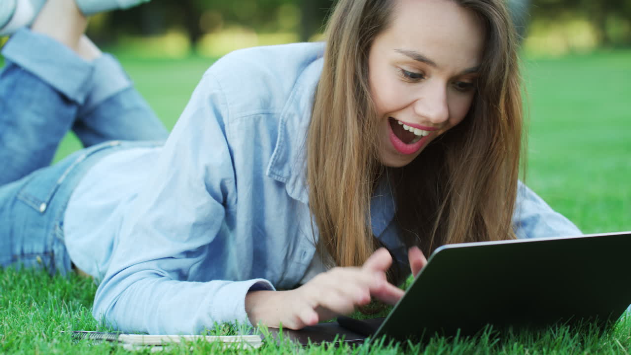 Smiling woman lying on green grass and typing on laptop in summer park
