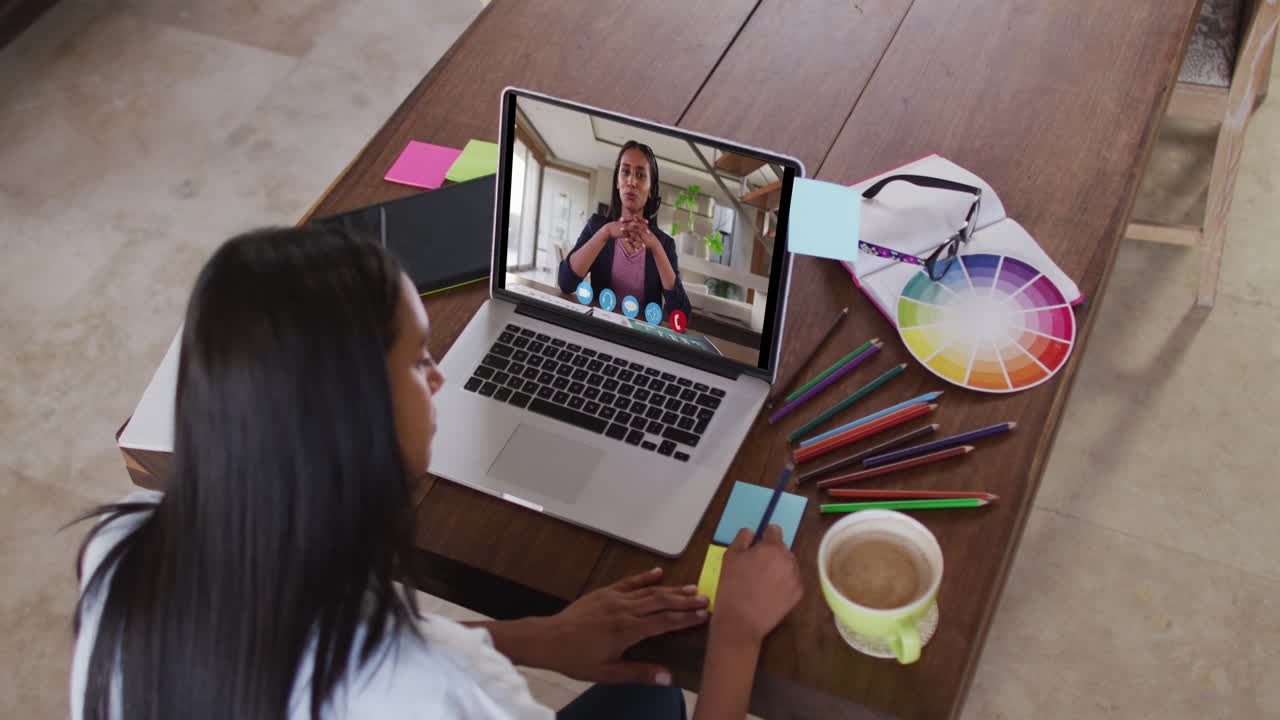 Mujer caucásica usando una laptop en videollamada con una colega femenina, tomando notas y trabajando desde casa