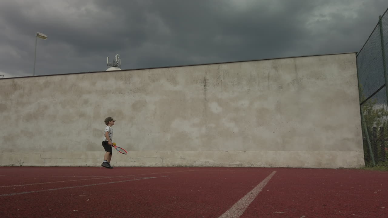 Little boy practices tennis against practice wall on court, low angle