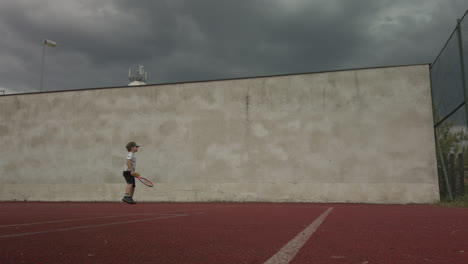 little boy practices tennis against practice wall on court, low angle