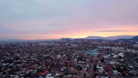 órbita-Aérea-De-La-Ciudad-De-Santiago-Con-Su-Sector-Financiero-Y-El-Atardecer-Entre-Montañas-Al-Fondo,-Chile