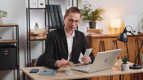 happy young caucasian businessman counting money cash and using laptop computer in home office