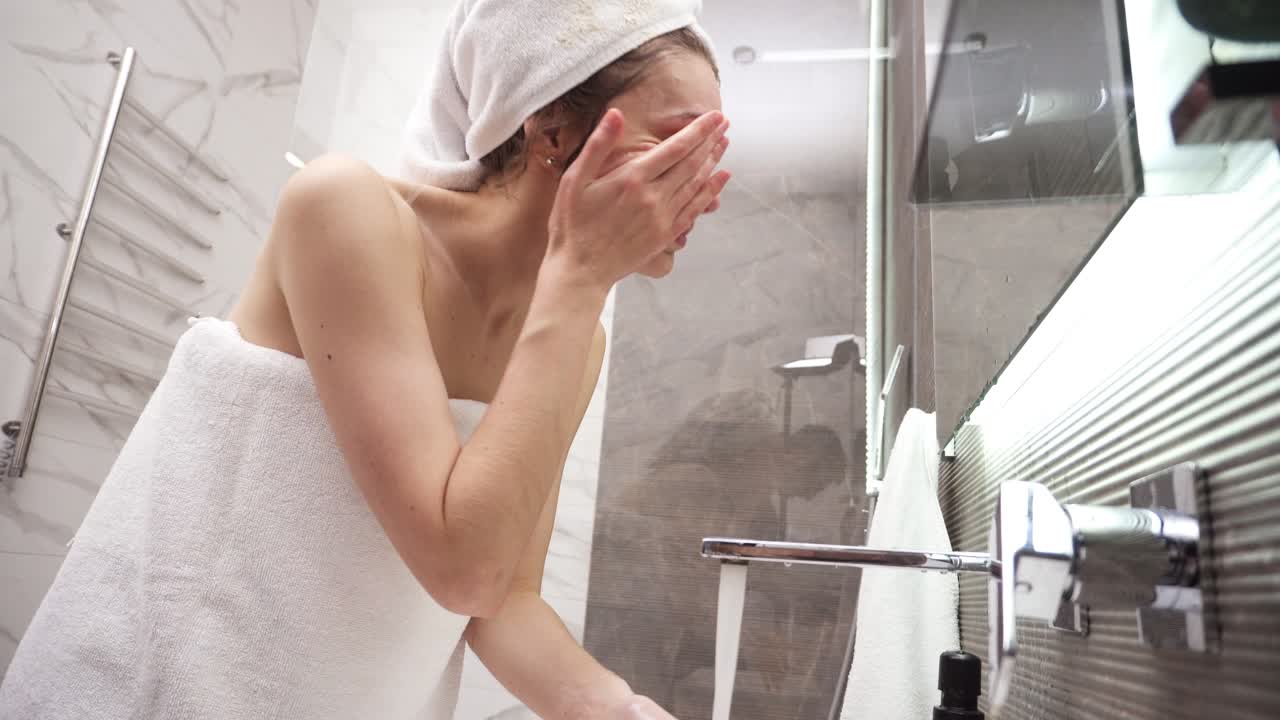 A woman standing in the bathroom near the sink in white towel on head and body washes her face with water from the tap. Looking to the mirror reflection and smiling. Low angle view