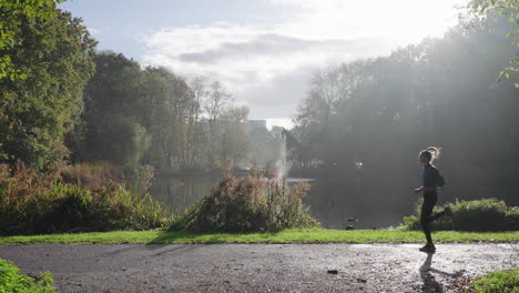 This-stock-footage-captures-a-woman-running-from-right-to-left-in-a-sunny-park-with-a-pond-in-the-background,-outdoor-exercise-amidst-beautiful-natural-surroundings