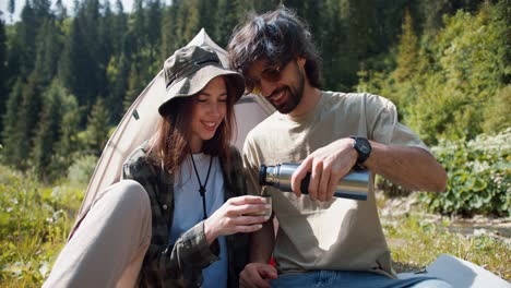 A-brunette-guy-in-yellow-sunglasses-pours-tea-from-a-thermos-to-a-brunette-girl-in-a-cap,-they-sit-near-the-tent-against-the-backdrop-of-a-green-forest