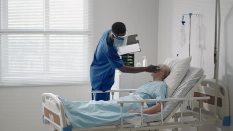 An-elderly-male-patient-lying-in-a-hospital-bed-connected-to-an-ECG-machine-talks-to-a-black-doctor-in-a-protective-mask