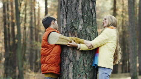 side view of caucasian sister and brother playing in the wood, hiding behind a tree trunk and looking at each other from different sides