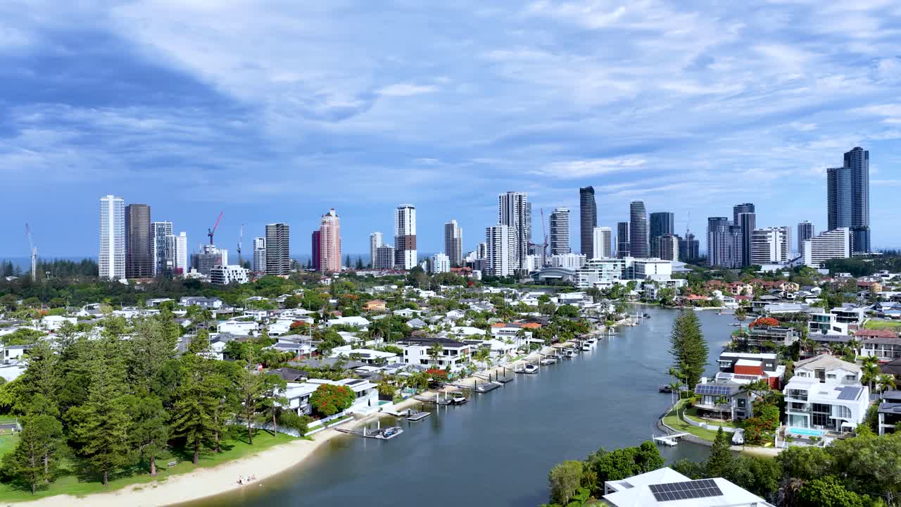 Drone glides above canal, luxury homes, private docks, pools, and city skyline in daylight