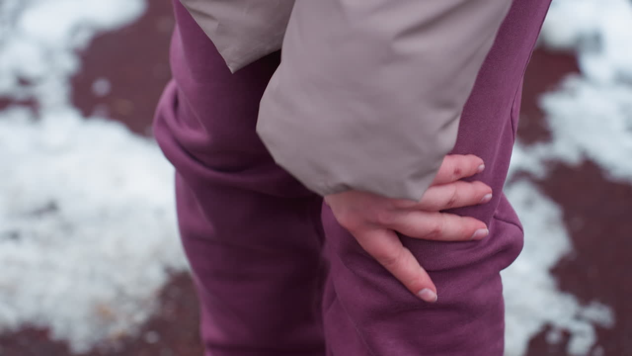 крупный план человека в зимнем наряде, потирающего левое колено, возможно, в боли, руки, нажимающие на коричневые брюки с видимым напряжением, размытый снежный фон