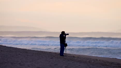 silueta de fotógrafo en la playa al amanecer con grandes olas borrosas en el fondo