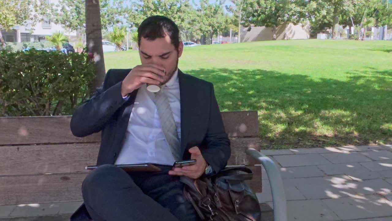 Forretningsmand der multitasker med kaffe, telefon og tablet i en park