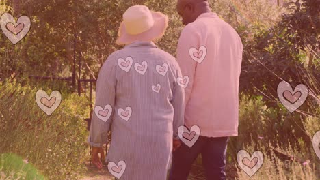 animation of hearts over senior african american couple walking in garden at christmas