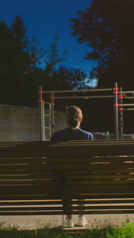 vista trasera de una mujer sentada en un banco al aire libre durante la noche, frente a una estructura de patio de recreo bajo una luz tenue, con un entorno sombreado, una pared en el fondo y un cielo azul profundo