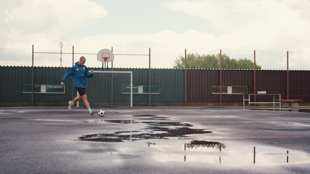 ensom fotballspiller på våt bane som dribler gjennom sølepytter under skyet himmel iført blå jakke slitt asfalt rustent gjerde mål ring bakgrunn reflekterende overflate rytmiske fotspor kornete urban