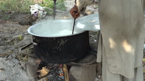 a man is carefully preparing milk in a large pot on a wooden stove, highlighting the purity of the natural environment.