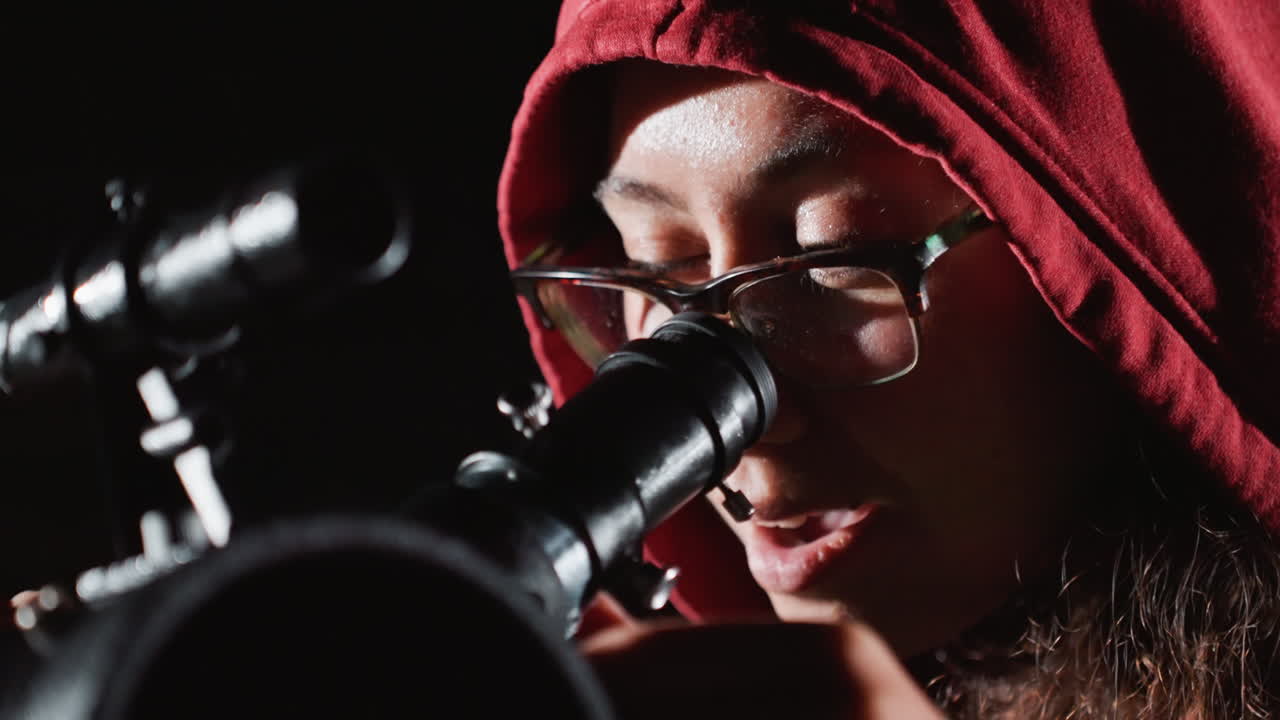 Studentessa con felpa bianca sul tetto che regola un telescopio, felpa con cappuccio rossa e occhiali, vista ravvicinata dell'oculare, mani che allineano il telescopio, improvviso sorriso dopo la scoperta, bokeh delle luci della città, espressione entusiasta, stupore giocoso