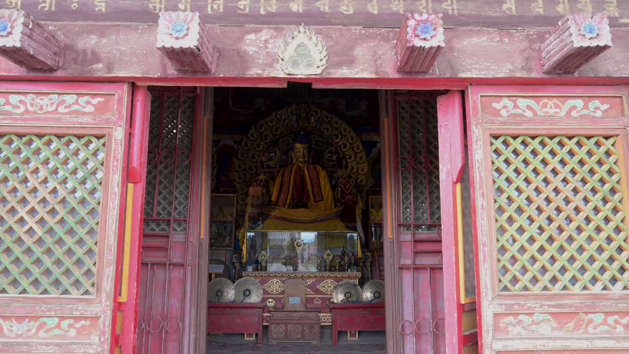 Vista attraverso elaborate porte a graticcio rosse del Monastero di Erdene Zuu, Mongolia: una statua dorata del Buddha è custodita all'interno di un padiglione storico con antichi manufatti sacri per l'altare e arte religiosa