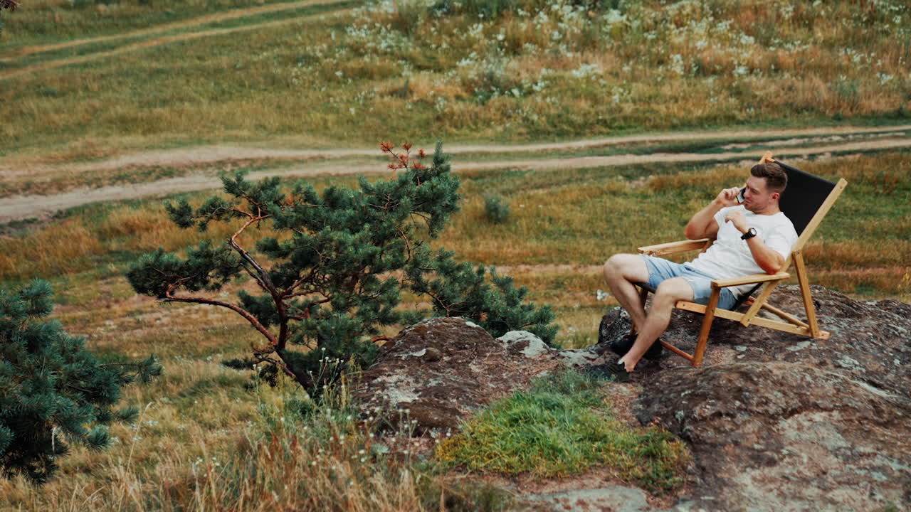 Mid-aged man sitting in the folding chair on the rock. Male entrepreneur speaks on the phone, working remotely in the nature.
