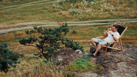 mid-aged man sitting in the folding chair on the rock. male entrepreneur speaks on the phone, working remotely in the nature.