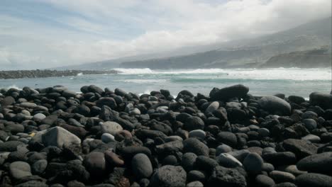 Slowmotion-group-of-surfers-revealed-behind-stones-on-pebble-beach,-Playa-Martiánez,-Puerto-de-la-Cruz,-Tenerife,-Canary-Islands-in-spring