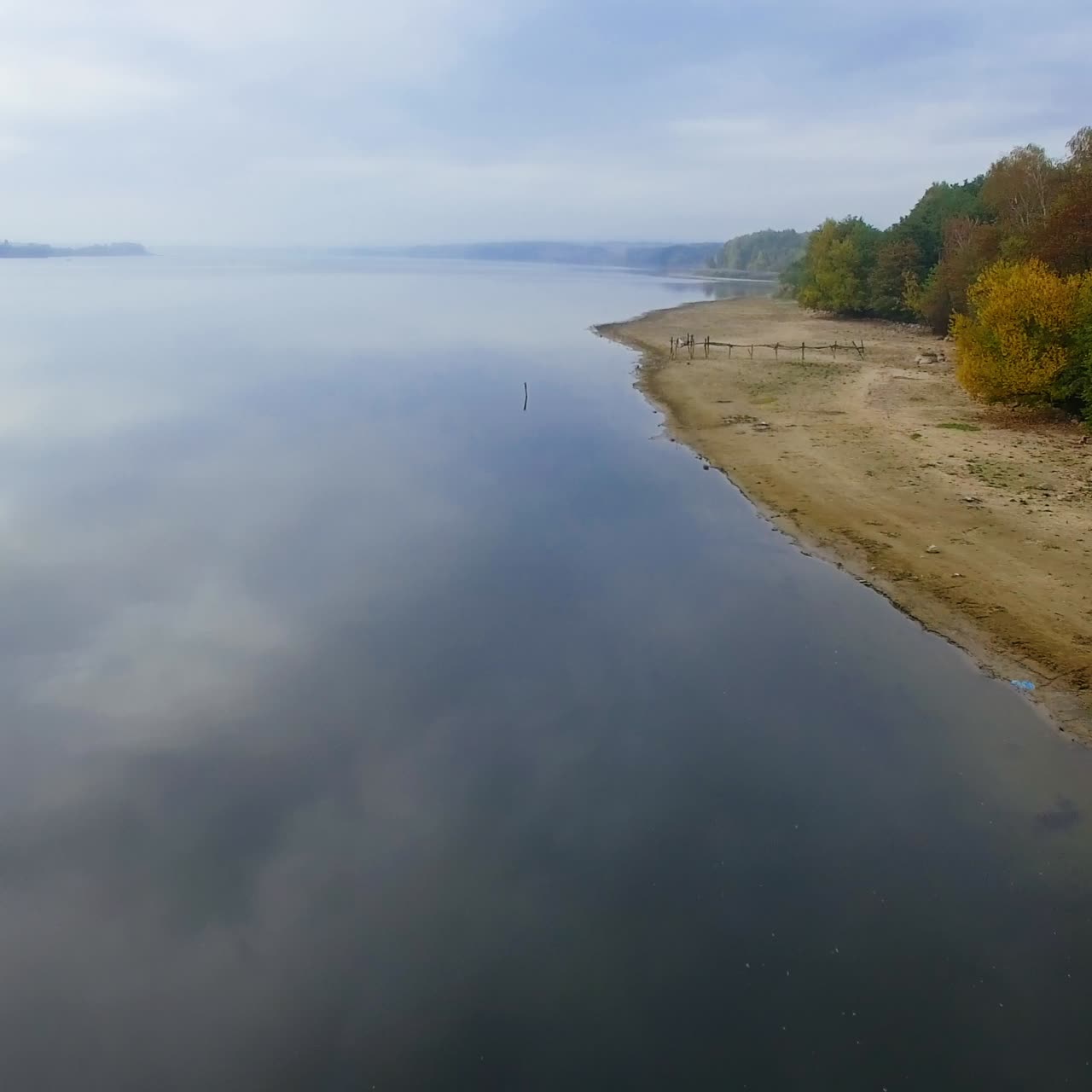 Dronevlucht boven het spiegelende oppervlak van de rivier op een bewolkte, grijze dag. Bovenaanzicht van de zanderige oever met bomen. Wazige horizon op de achtergrond