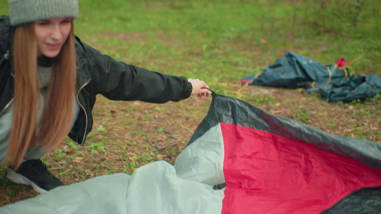 Close up of young lady wearing hoodie and black jacket holding edge of tent fabric while setting up camp in forest area, adjusting position on grassy ground surrounded by trees
