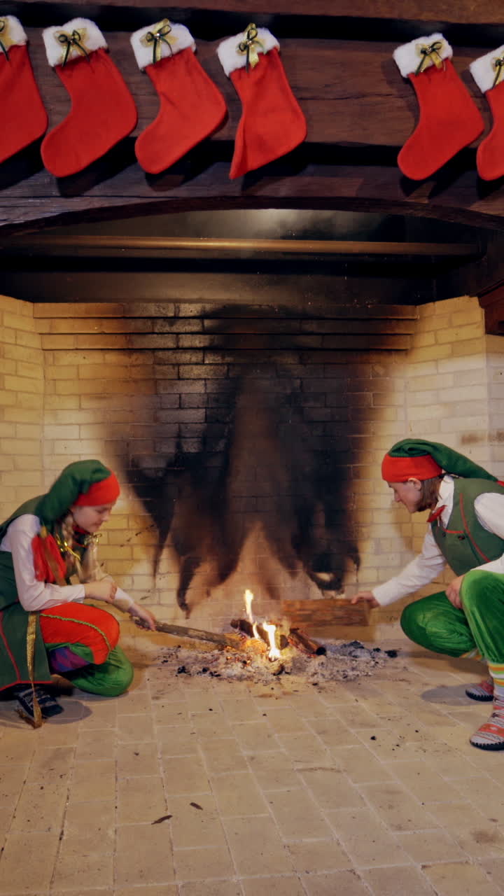 Elves near fireplace on Christmas background indoors. Funny elves in green costumes sit near the fire in the house. Vertical video