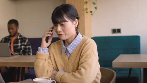 Mujer-Hablando-Por-Teléfono-Celular-Sentada-En-Una-Mesa-En-Una-Cafetería-Mientras-Un-Hombre-Trabaja-En-Una-Computadora-Portátil-Detrás-De-Ella