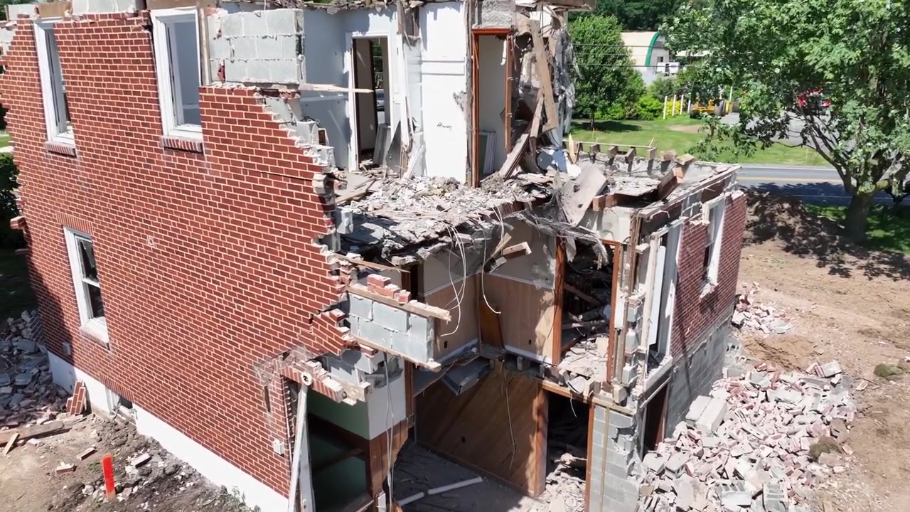 Escavadeira demolindo casa. Demolição ao nível do solo com detritos caindo. Vista aérea de casa condenada sendo demolida. Dia ensolarado em subúrbio americano. Parede de casa destruída.