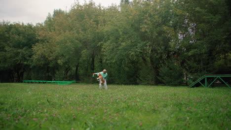 dog owner playing fetch with dog on grassy field surrounded by tall trees, throwing ball for dog to chase while cars pass by in distance under cloudy sky, creating joyful active outdoor scene