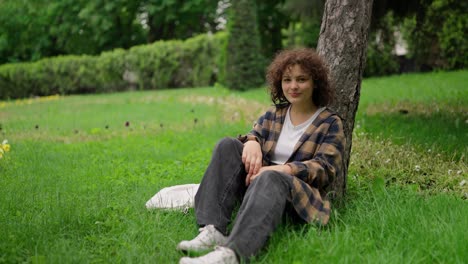 Portrait-of-a-happy-brunette-girl-in-a-plaid-shirt-who-is-sitting-near-a-tree-and-posing-on-the-grass-in-the-park