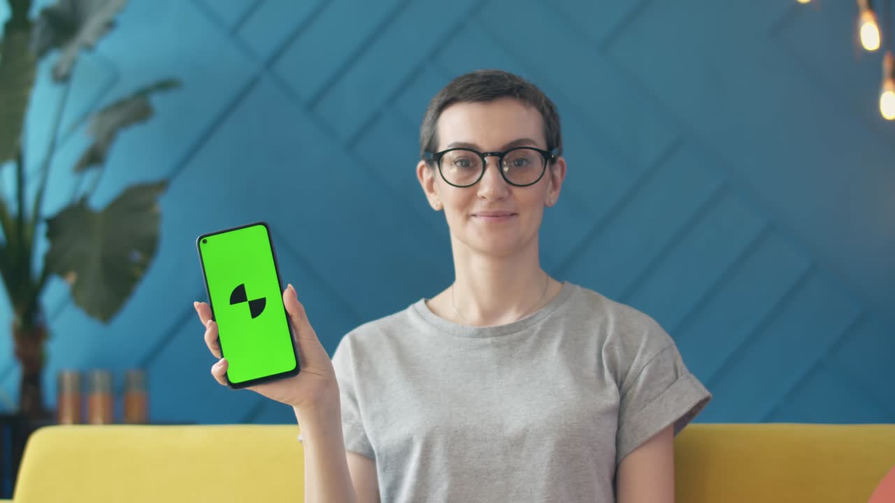mujer linda con gafas mirando a la cámara y sentada en el sofá sosteniendo un teléfono con una aplicación en la mano, pantalla verde. concepto de publicidad en línea y aplicaciones