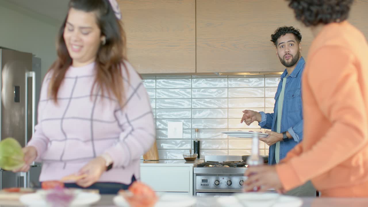 Diversi amici gesticolano offrendo bibite, mentre una donna vivace affetta lattuga su un tagliere in cucina per un pasto.