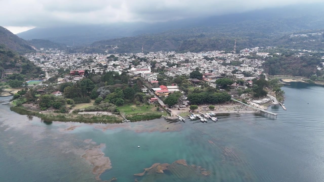 Ripresa aerea con drone su San Lucas, Guatemala, rivela acque scintillanti, quartieri tropicali, palme e una catena vulcanica circostante con cime boscose, mostrando un vivido paesaggio centroamericano.