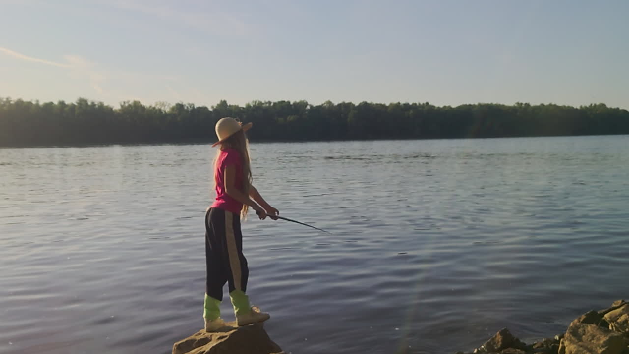 Sebuah bidikan statis lebar profesional menangkap seorang anak mengenakan topi matahari saat memancing dari tepi berbatu Danube. Cahaya sore yang lembut menerangi air yang tenang dan deretan pohon di kejauhan