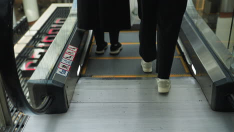 close-up view of two people in black outfits ascending an escalator in a busy mall. the focus is on their movement and the details of the escalator steps