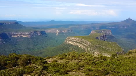 Drone-flying-over-tree-lined-cliff-edge-revealing-deep-valley,-mountains-and-rocky-escarpments