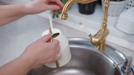 Character with plaid shirt fetches water from modern brass faucet over stainless sink into sleek white kettle then shuts tap off amid lush indoor potted greenery on cozy kitchen counter